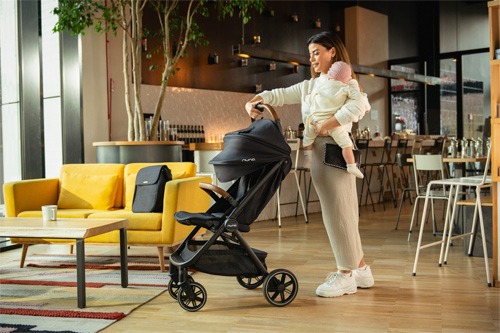 madre con hijo en brazos plegando la silla de paseo TRVL LX de Nuna en una cafetería